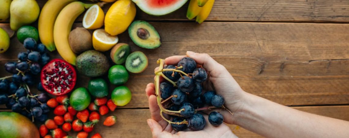 A variety of fresh, organic fruits with hands holding grapes on a wooden table.