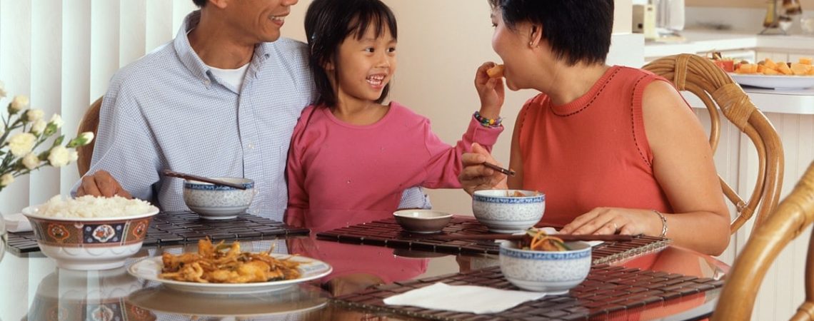 An Asian family, an adult male and female are seated around a table eating a meal with a young female standing in between the adults.